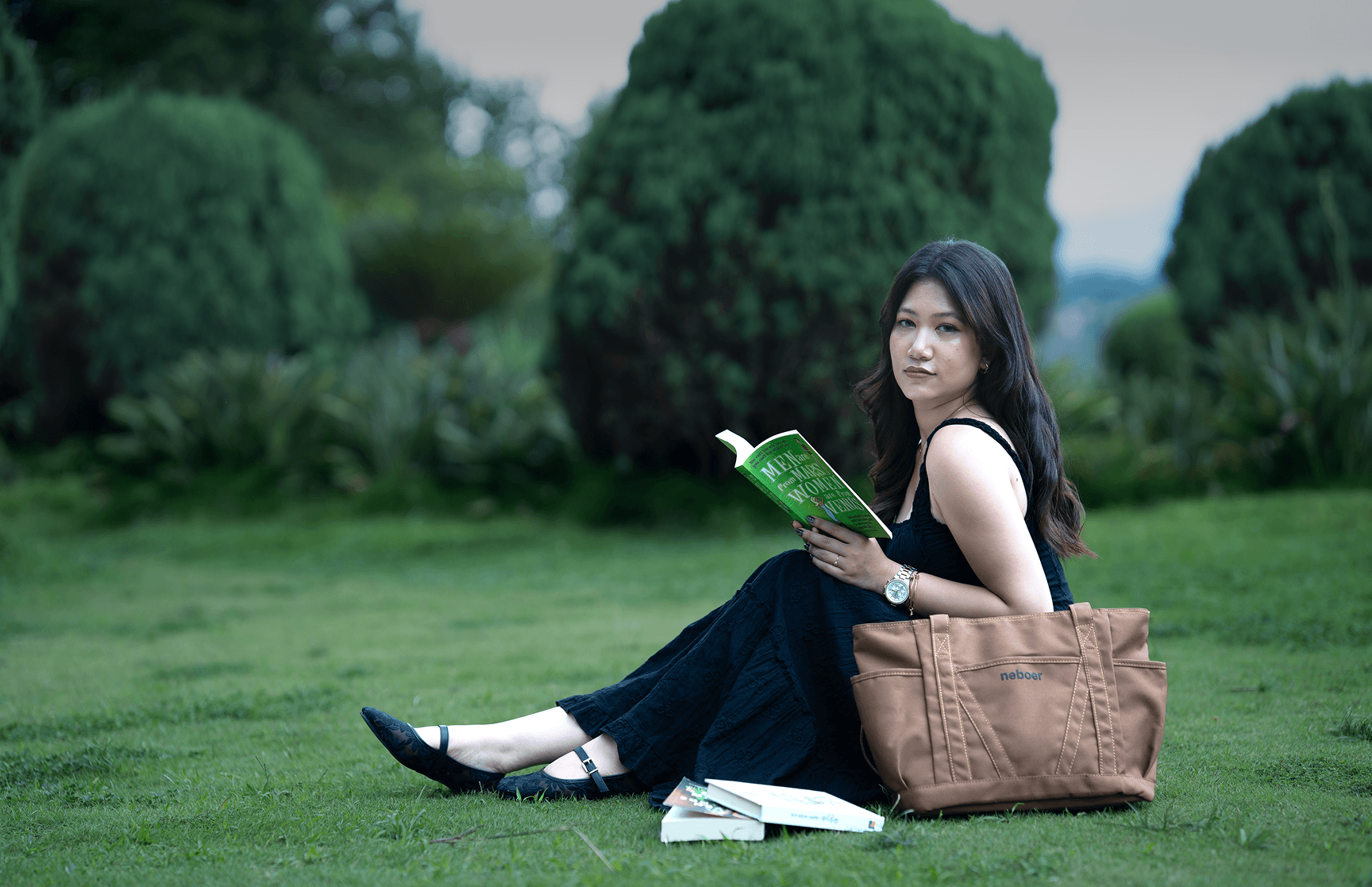 Woman sitting on grass reading, with a Neboer tote bag